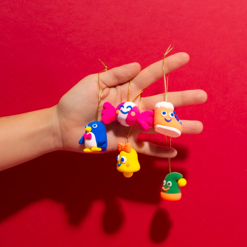 Hand hält 5 Weihnachtsdekorationen aus OMY Modelliermasse: Pinguin, Zuckerstange, Stiefel, Glocke und Mütze, auf rotem Hintergrund.