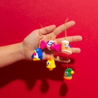 Hand hält 5 Weihnachtsdekorationen aus OMY Modelliermasse: Pinguin, Zuckerstange, Stiefel, Glocke und Mütze, auf rotem Hintergrund.