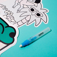 Gros plan sur le set de table Magic Set Dino OMY à colorier et son feutre bleu OMY, sur fond turquoise