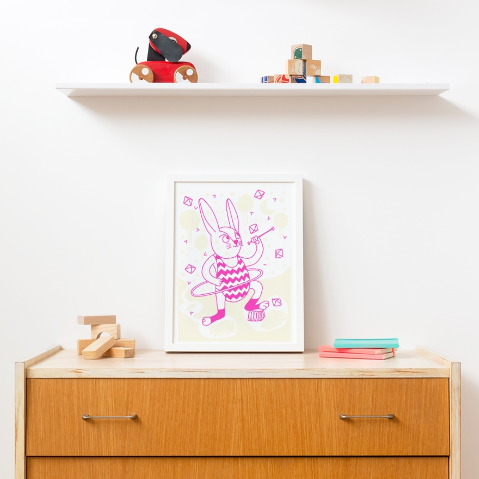 Kinderzimmer: gerahmtes OMY Poster rosa Hase mit Hula-Hoop-Reifen auf einer Holzkommode mit Spielzeug und Büchern