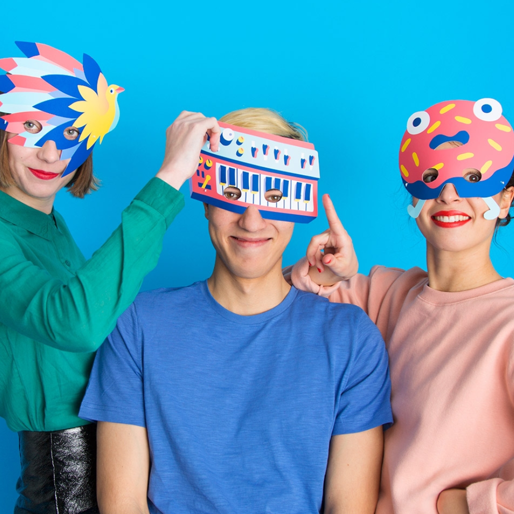 Drei lächelnde Freunde tragen die OMY Masks Party Masken: Vogel, Synthesizer und Donut, auf blauem Hintergrund