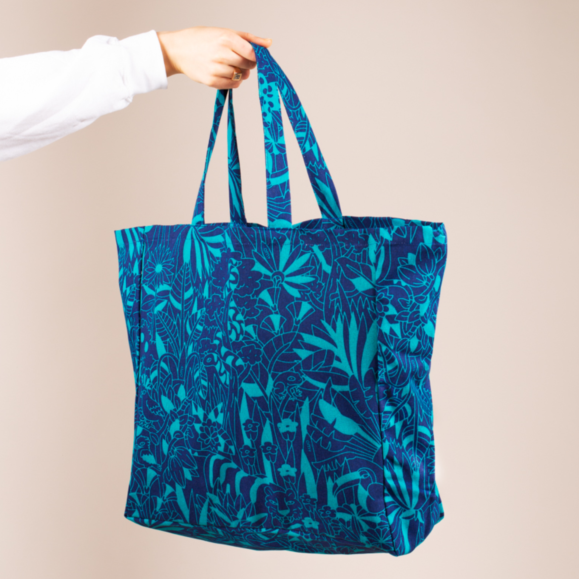 Main tenant un sac cabas OMY en toile imprimée jungle bleu marine et turquoise, sur fond rose pâle
