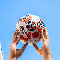 Mains levant vers le ciel bleu un ballon de foot OMY illustré de motifs orange et noirs : tigre, soleil, ballon souriant