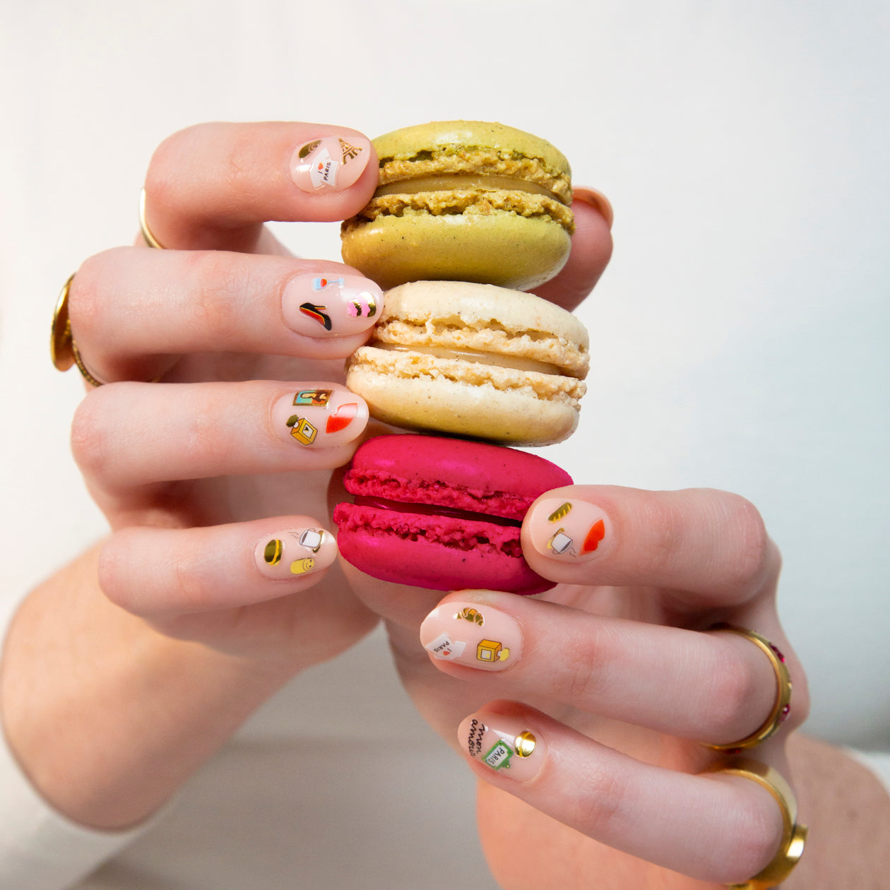 Handen met nagels versierd met Paris OMY-nagelstickers, houden een stapel macarons met pistache, vanille en framboos vast.