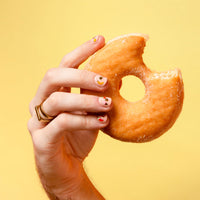 Main aux ongles décorés de stickers pour ongles Yummy OMY tenant un donut entamé, sur fond jaune
