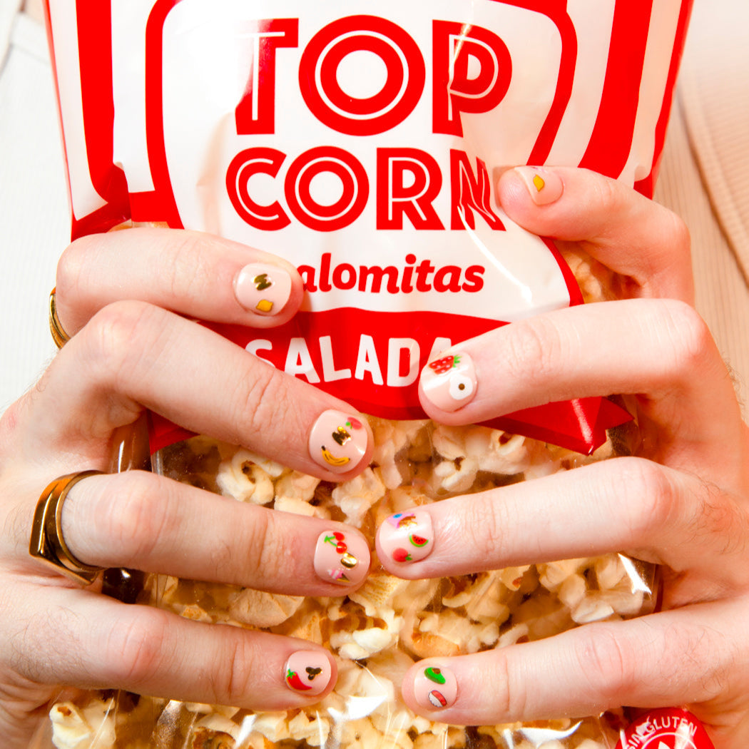 Mains aux ongles décorés de stickers pour ongles Yummy OMY tenant un sachet de pop-corn rouge et blanc
