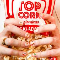 Mains aux ongles décorés de stickers pour ongles Yummy OMY tenant un sachet de pop-corn rouge et blanc