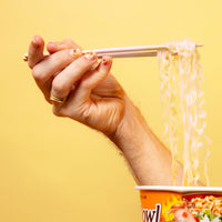 Main aux ongles décorés de stickers pour ongles Yummy OMY soulevant des nouilles avec des baguettes, sur fond jaune