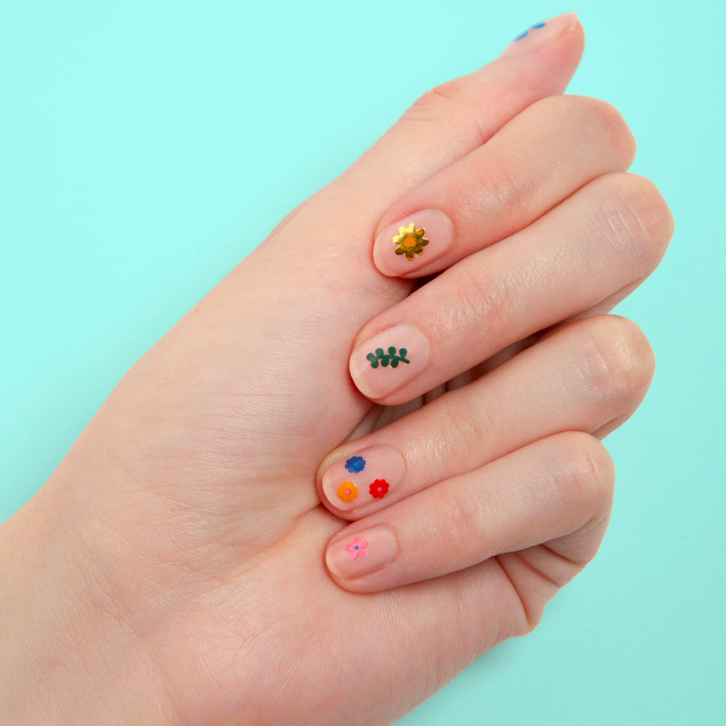Hand met nude nagels versierd met Flower OMY-nagelstickers: kleurrijke bloemen, een gouden bloem en loof, op een turquoise achtergrond.