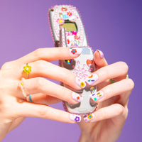 Handen met nagels versierd met Flower OMY-nagelstickers, houden een klaptelefoon met strass vast, op een paarse achtergrond.
