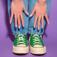 Handen met nagels versierd met Flower OMY-nagelstickers op een spijkerbroek, groene sneakers, op een paarse achtergrond.