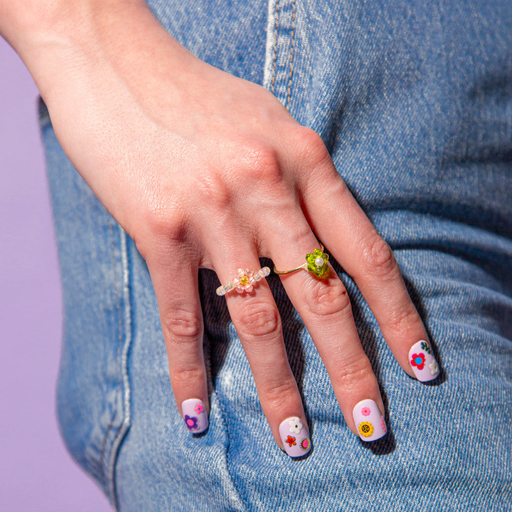 Hand op een spijkerbroek, lila nagels versierd met OMY Flower Nail Stickers: kleine kleurrijke zelfklevende bloemetjes.