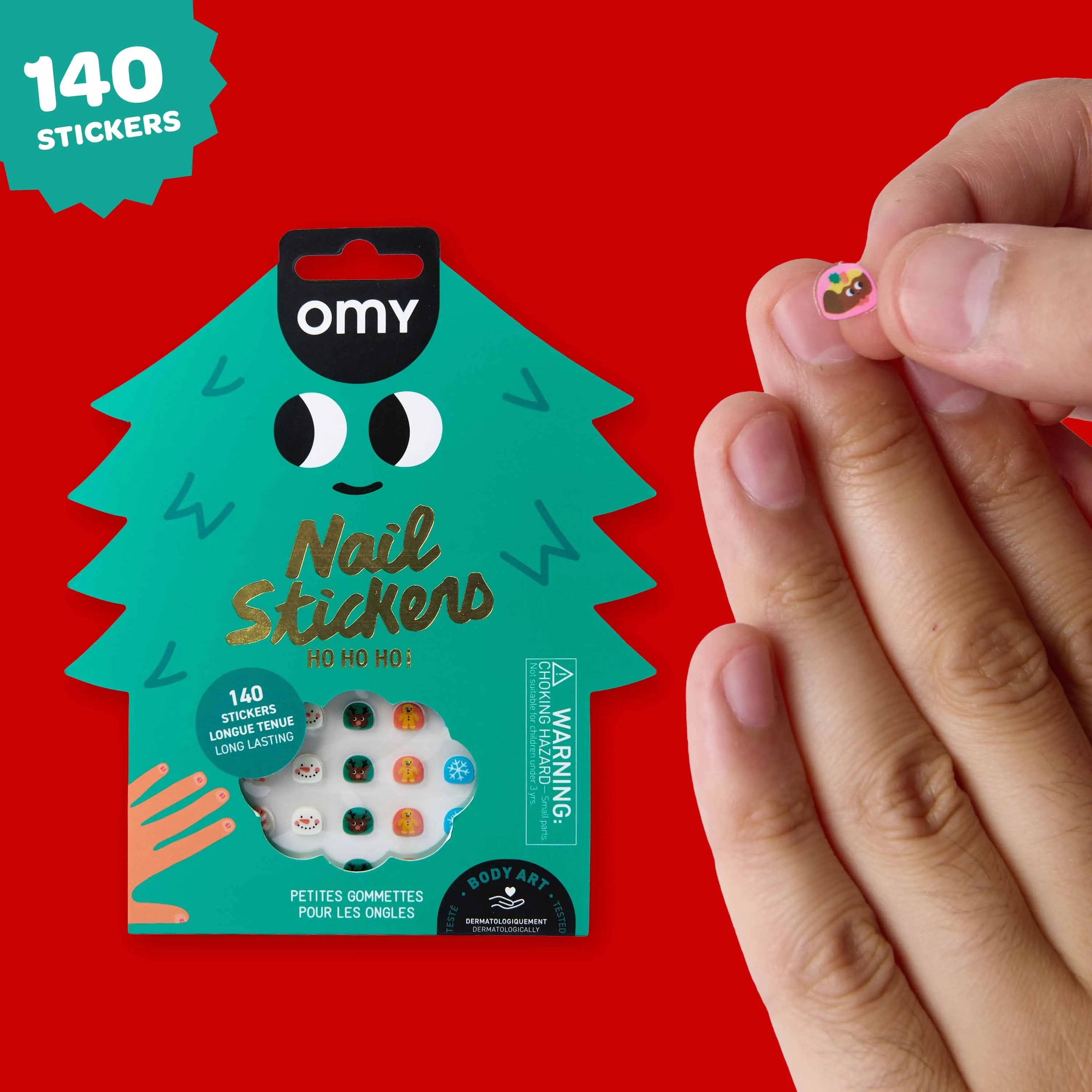 Pochette de 140 nail stickers OMY aux motifs de Noël, avec un sticker présenté entre les doigts.