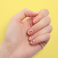 Main aux ongles nude décorés de stickers pour ongles Yummy OMY : banane, frites, onigiri et pastèque, sur fond jaune