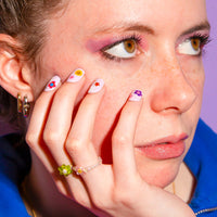 Nadenkende vrouw, hand tegen de wang, witte nagels versierd met Flower OMY-nagelstickers en ringen van bloemkralen.