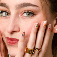 Lachende vrouw, hand tegen de wang, nude nagels versierd met Paris OMY-nagelstickers en gouden ringen.