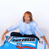 Jovem sentado de pernas cruzadas a brincar com o pufe insuflável OMY Fresh em forma de tubo de dentífrico azul, sobre fundo branco.