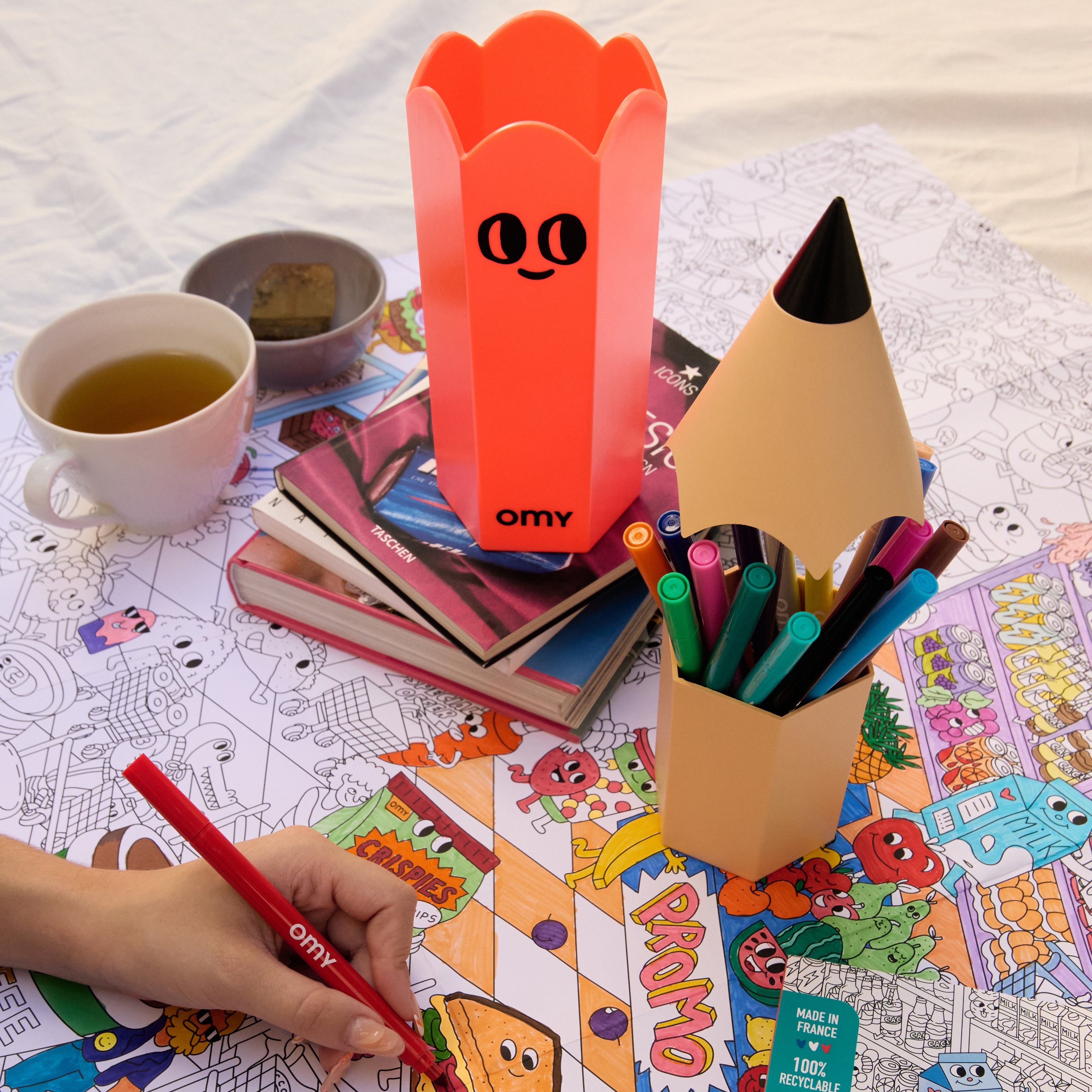 Set créatif OMY avec boîte à crayons ouverte remplie de feutres colorés, posée sur une table de coloriage en pleine activité avec tasse de thé et livres.