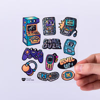 Planche de stickers Game Boy OMY avec consoles rétro, manettes, bornes d’arcade et messages pixelisés holographiques