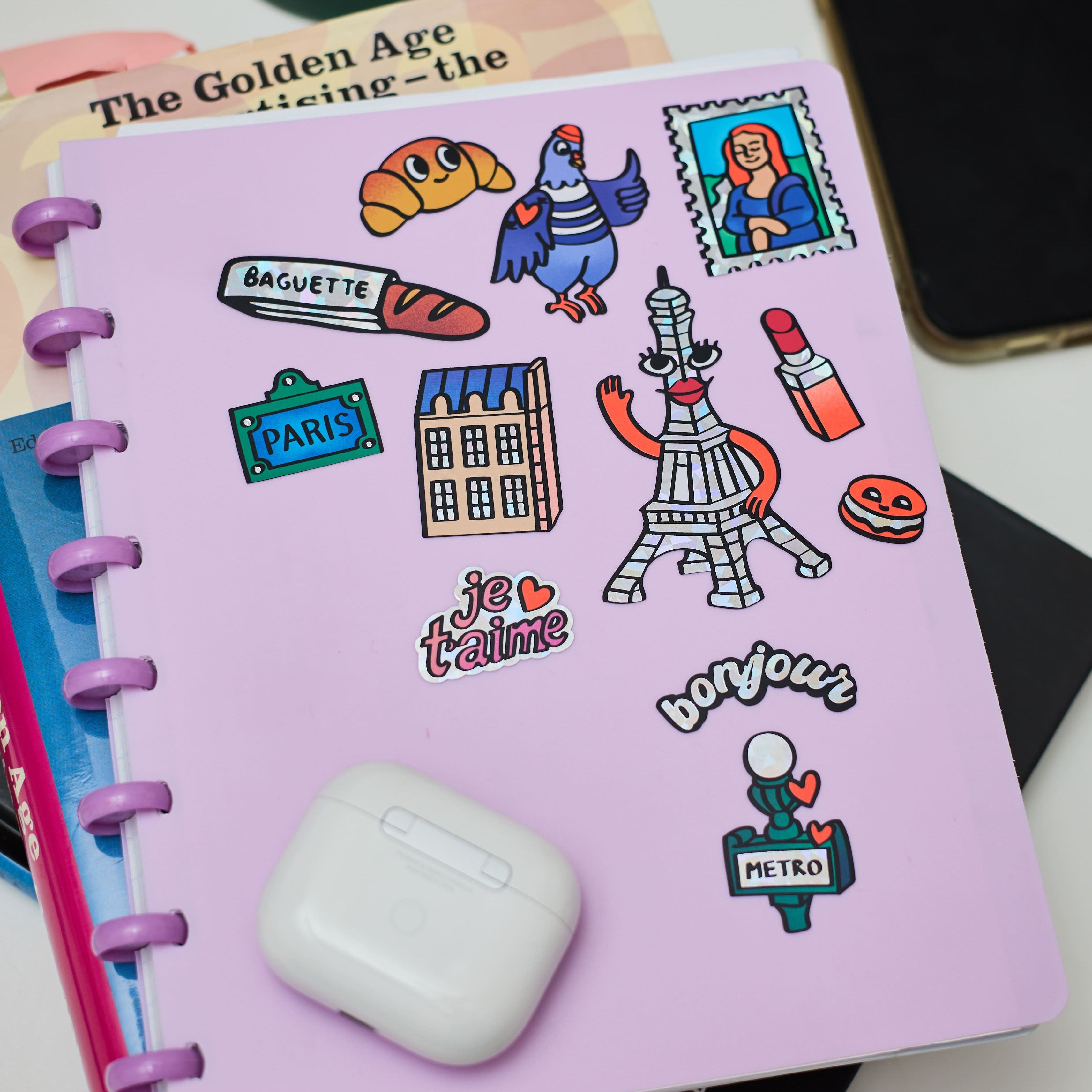 Stickers Paris holographiques collés sur un carnet violet : Tour Eiffel, baguette, pigeon, macarons, métro parisien, texte “je t’aime” et “bonjour”.