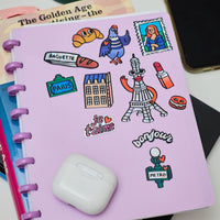 Stickers Paris holographiques collés sur un carnet violet : Tour Eiffel, baguette, pigeon, macarons, métro parisien, texte “je t’aime” et “bonjour”.