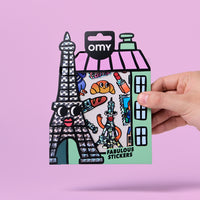 Planche de stickers Paris OMY avec Tour Eiffel holographique, illustrations parisiennes colorées et packaging en forme de boutique, tenue à la main sur fond rose.