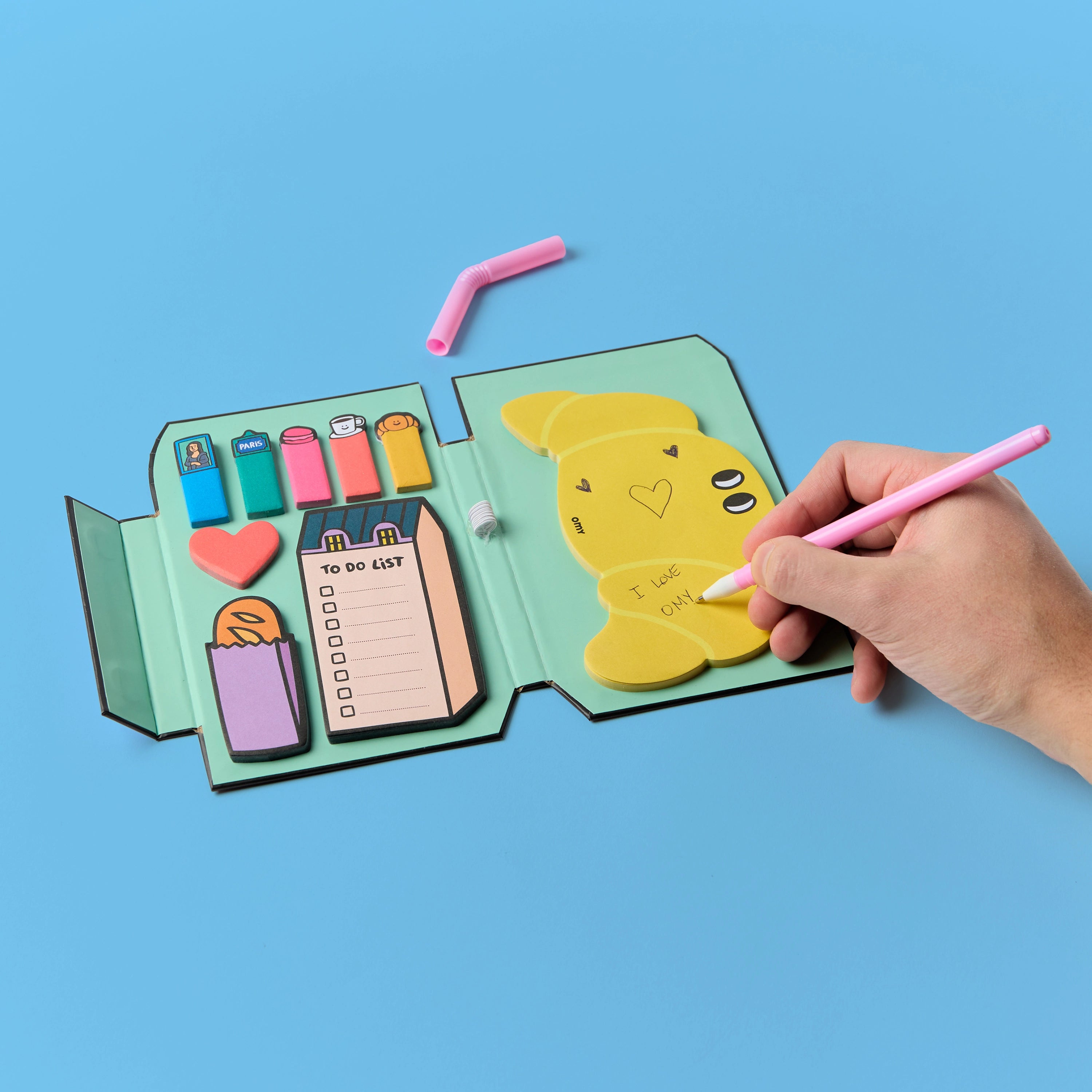 Sticky Notes Paris OMY ouvert – Mémos repositionnables, to-do list et notes autocollantes, une main écrit sur la page, fond bleu
