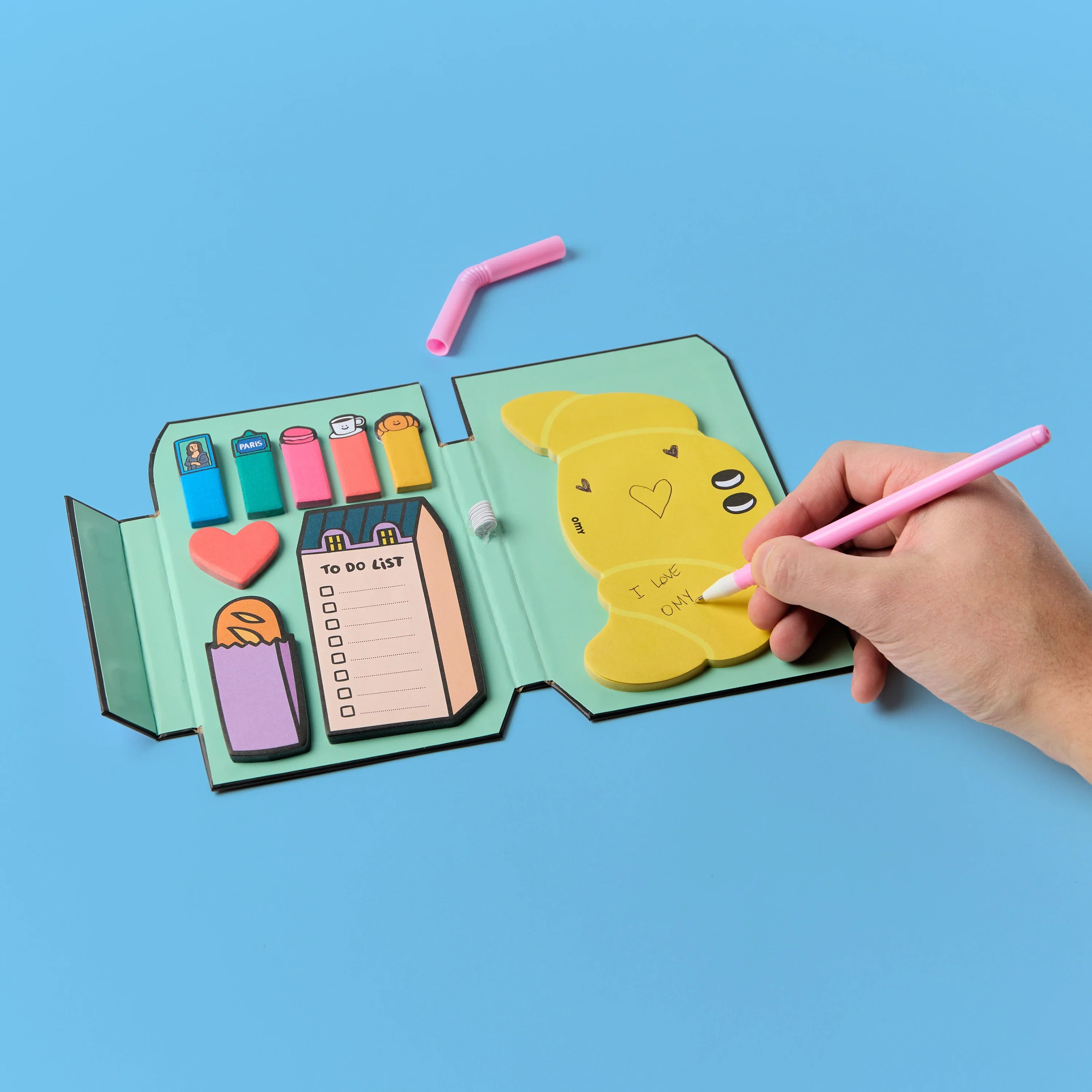 Sticky Notes Paris OMY ouvert – Mémos repositionnables, to-do list et notes autocollantes, une main écrit sur la page, fond bleu