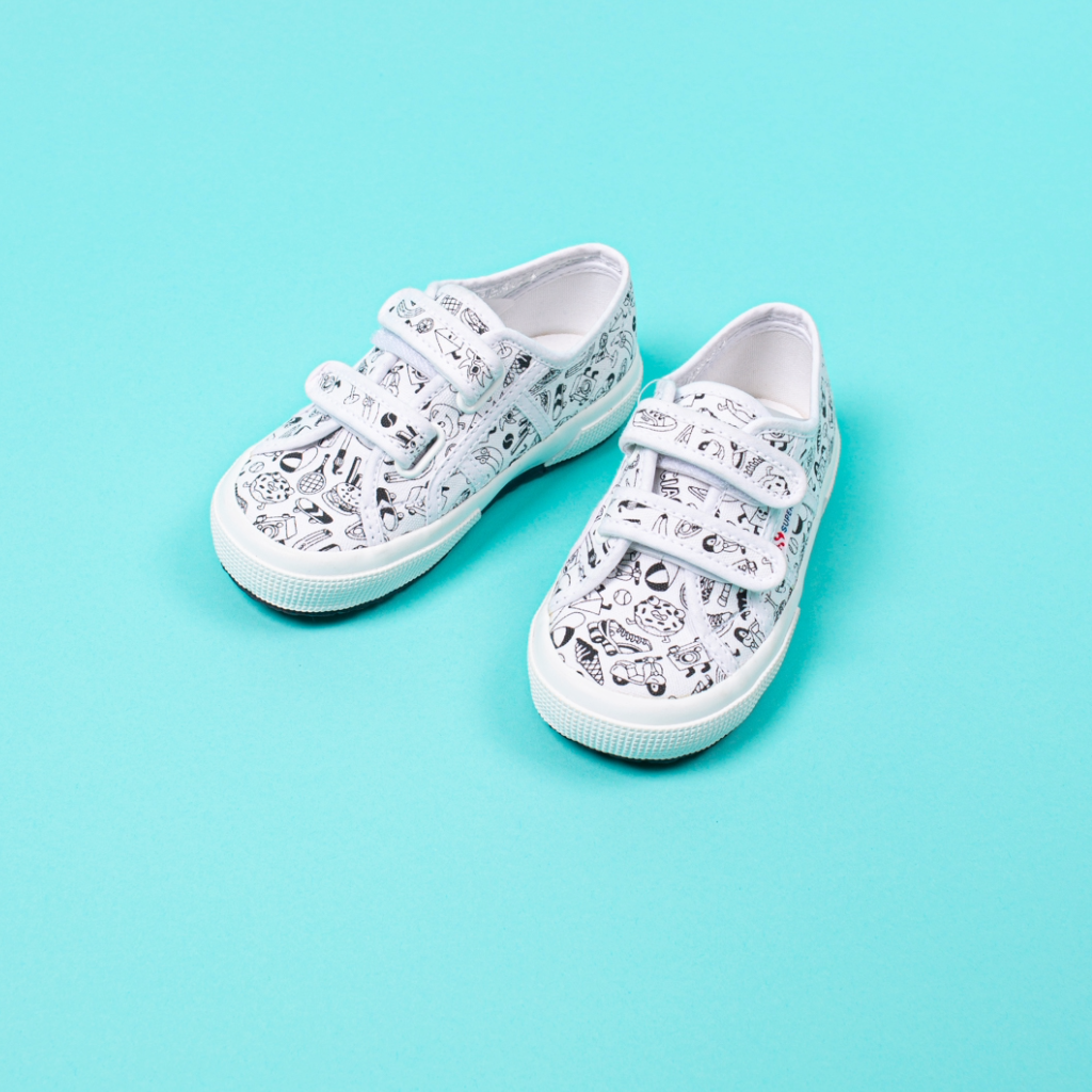 Pair of kids' Superga x OMY velcro sneakers with black-and-white illustrations to color, on a turquoise background