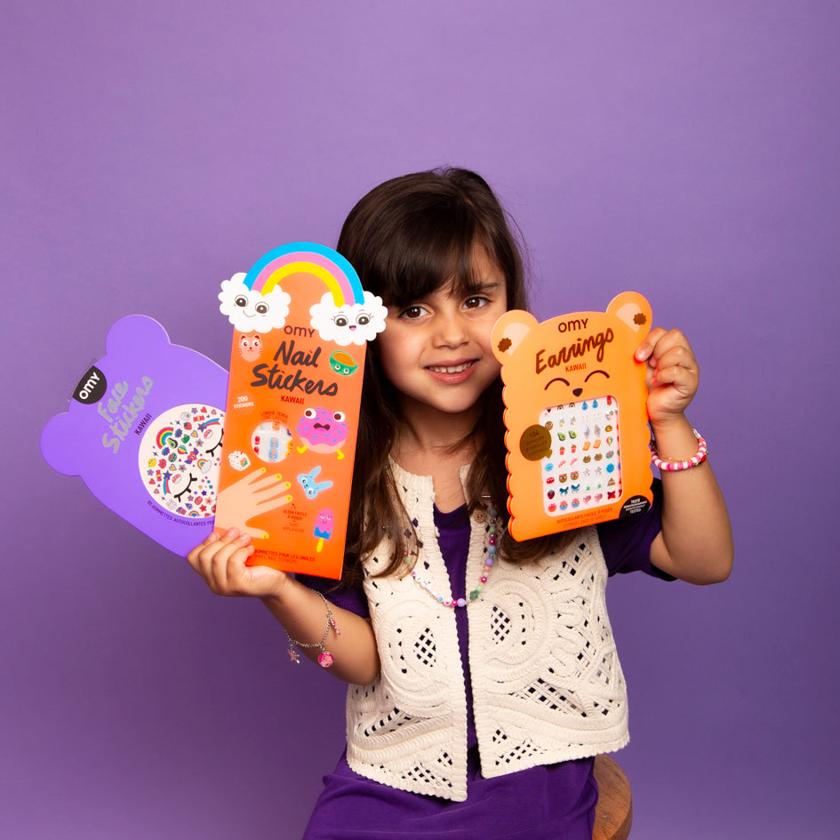 Fillette souriante tenant les Face Stickers, Nail Stickers et Earrings Kawaii OMY sur fond violet