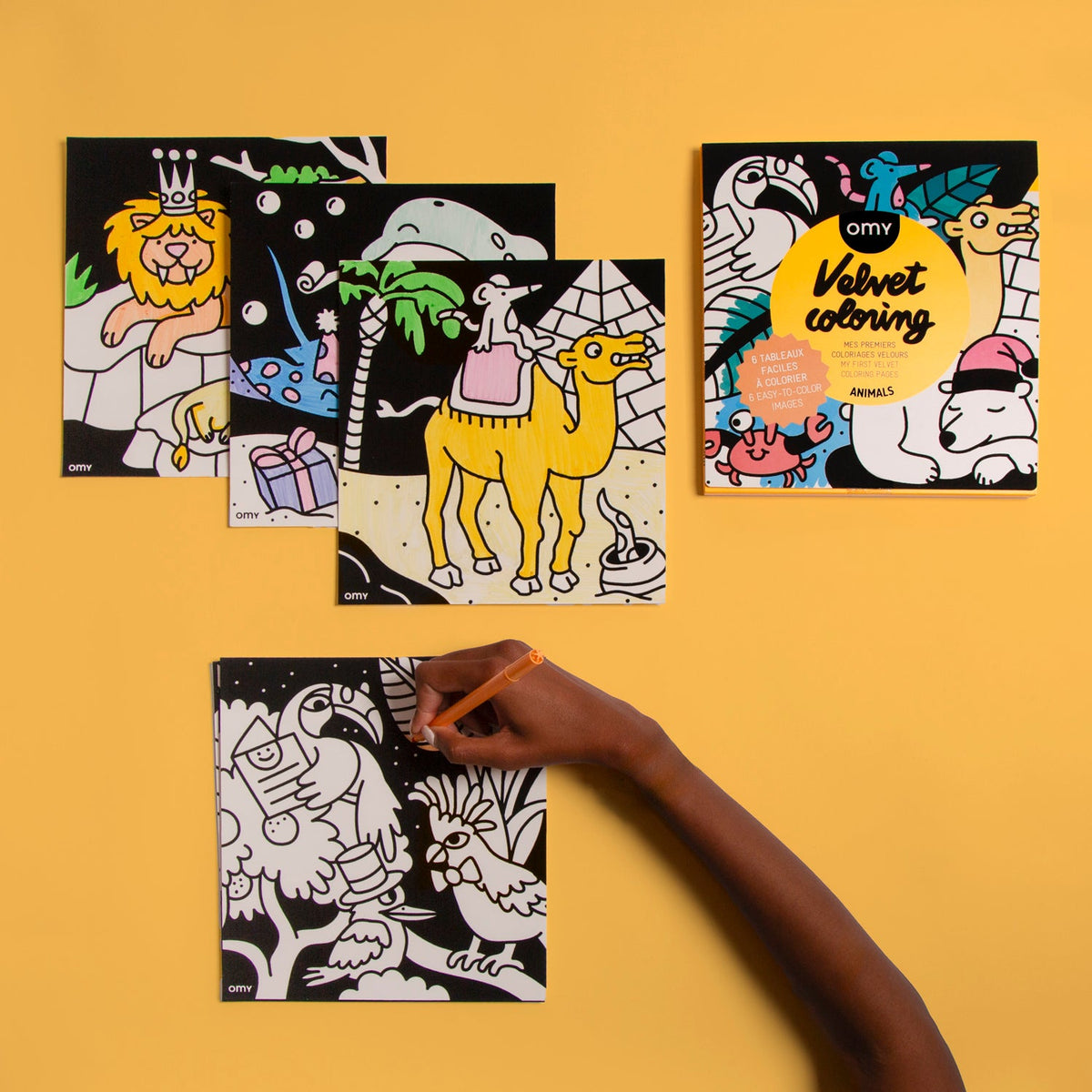 Velvet Coloring Animals OMY packaging with colored velvet pictures of a lion, dolphin and camel, and a hand coloring toucans, on a yellow background