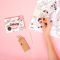 Hand malt das OMY Princess & Dragon Coloring Pocket Poster mit Buntstiften aus, neben seiner Box, auf rosa Hintergrund