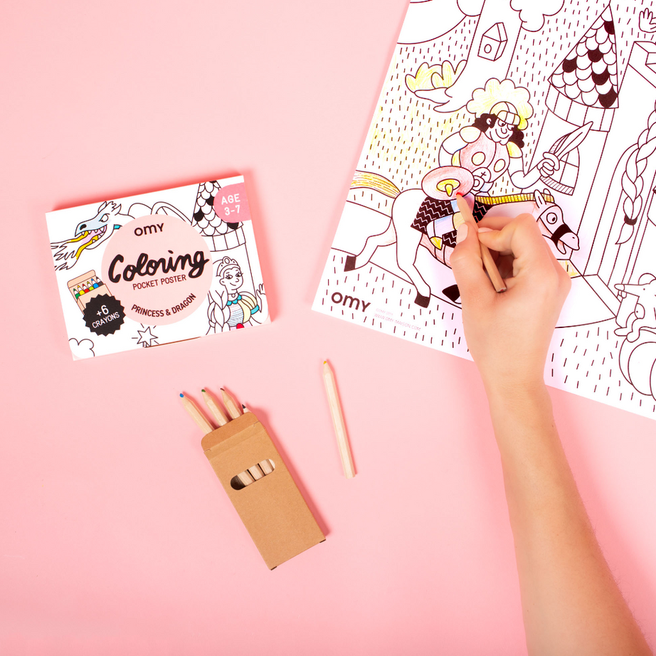 Mano che colora il Coloring Pocket Poster OMY Princess & Dragon con matite colorate, accanto alla sua scatola, su sfondo rosa