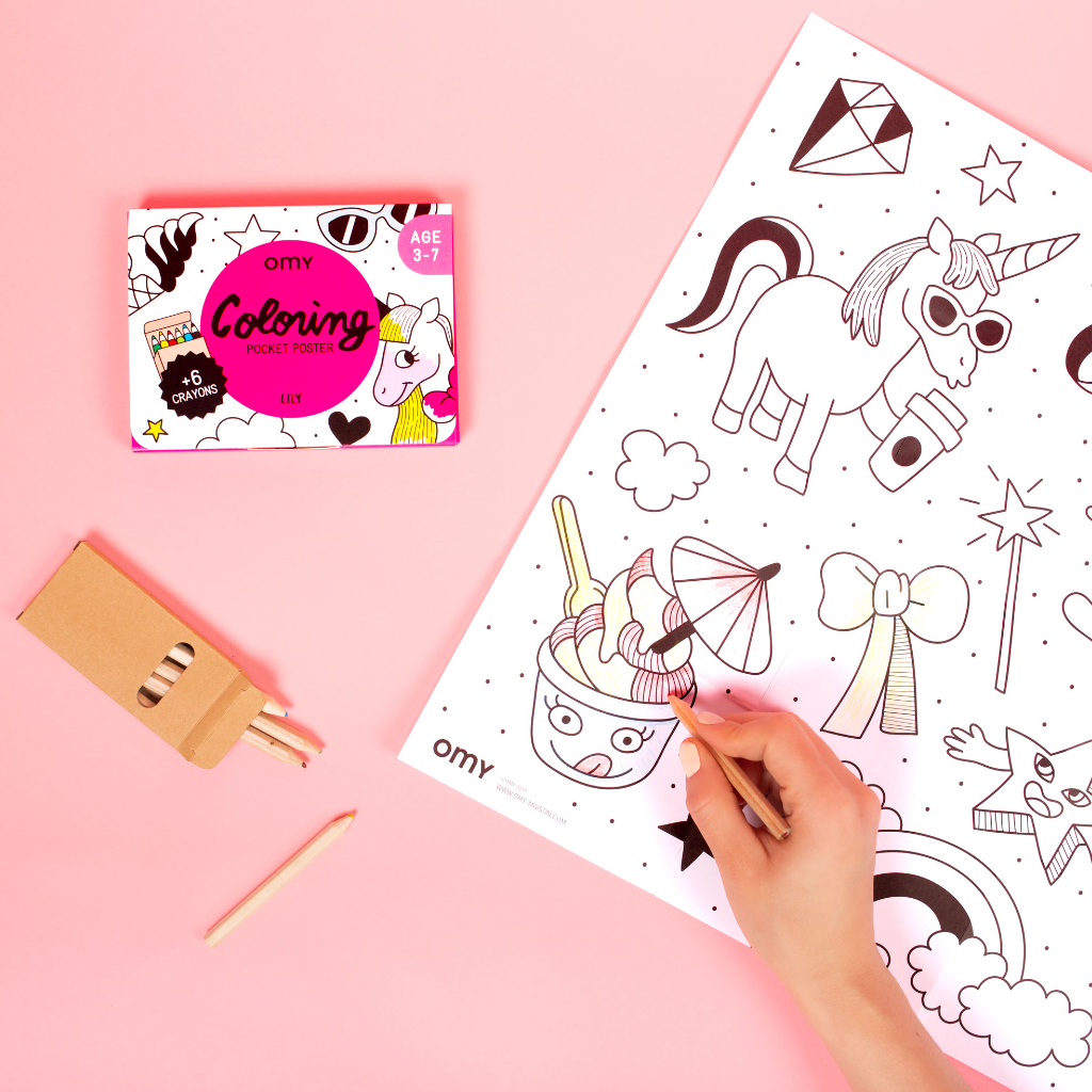 Mano coloreando el Coloring Pocket Poster OMY Lily con unicornios con lápices, junto a su caja rosa, sobre fondo rosa