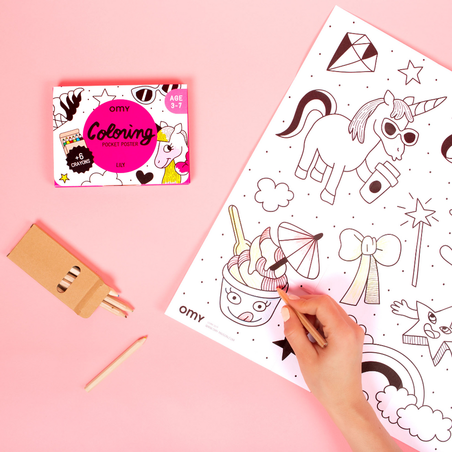 Main coloriant le Coloring Pocket Poster Lily OMY aux licornes avec des crayons, à côté de sa boîte rose, sur fond rose
